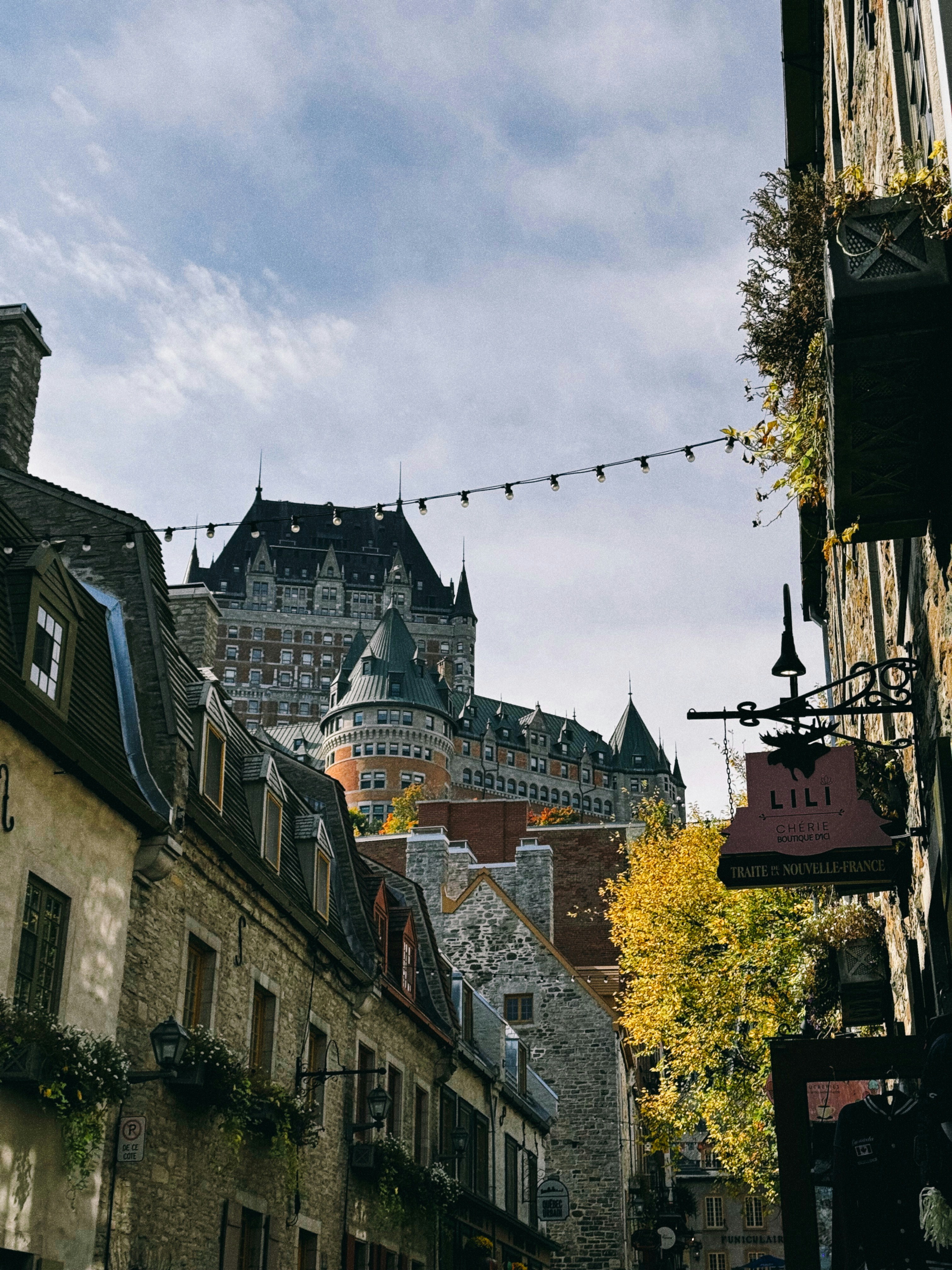 A narrow cobblestone street in Old Quebec strung with lights, the towers of the Chateau Frontenac rising above the rooftops in fall