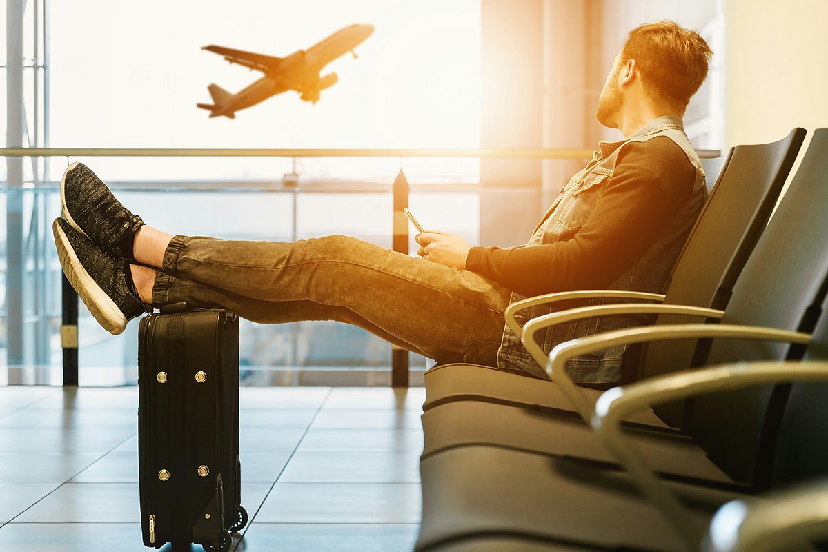 A traveler with his feet up on a suitcase watches a plane take off through an airport window