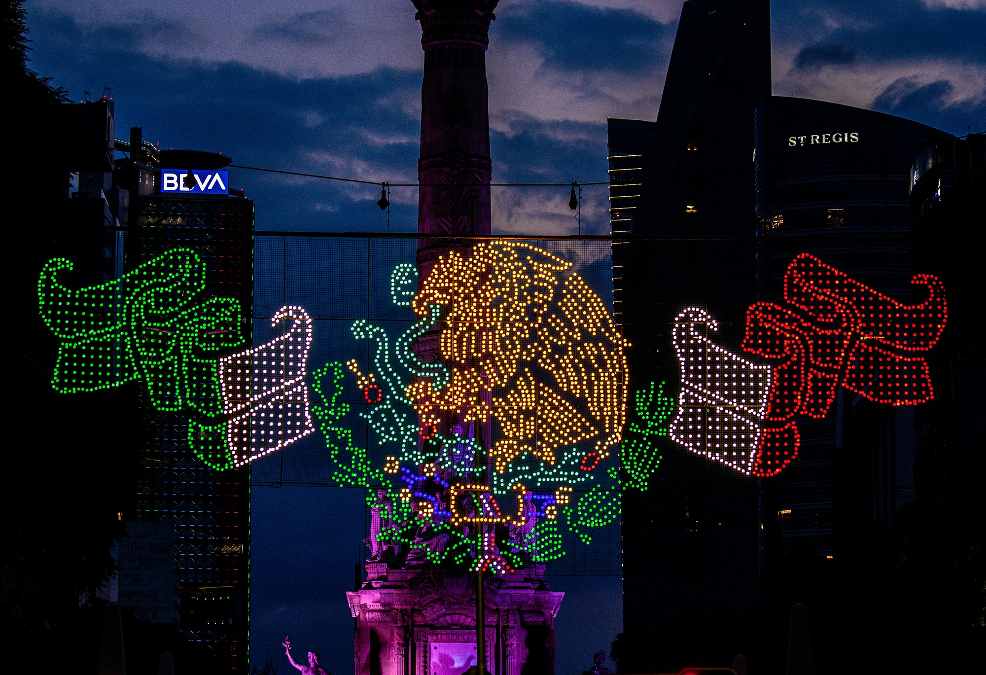 An illuminated light display of the Mexican coat of arms above Paseo de la Reforma in Mexico City at dusk