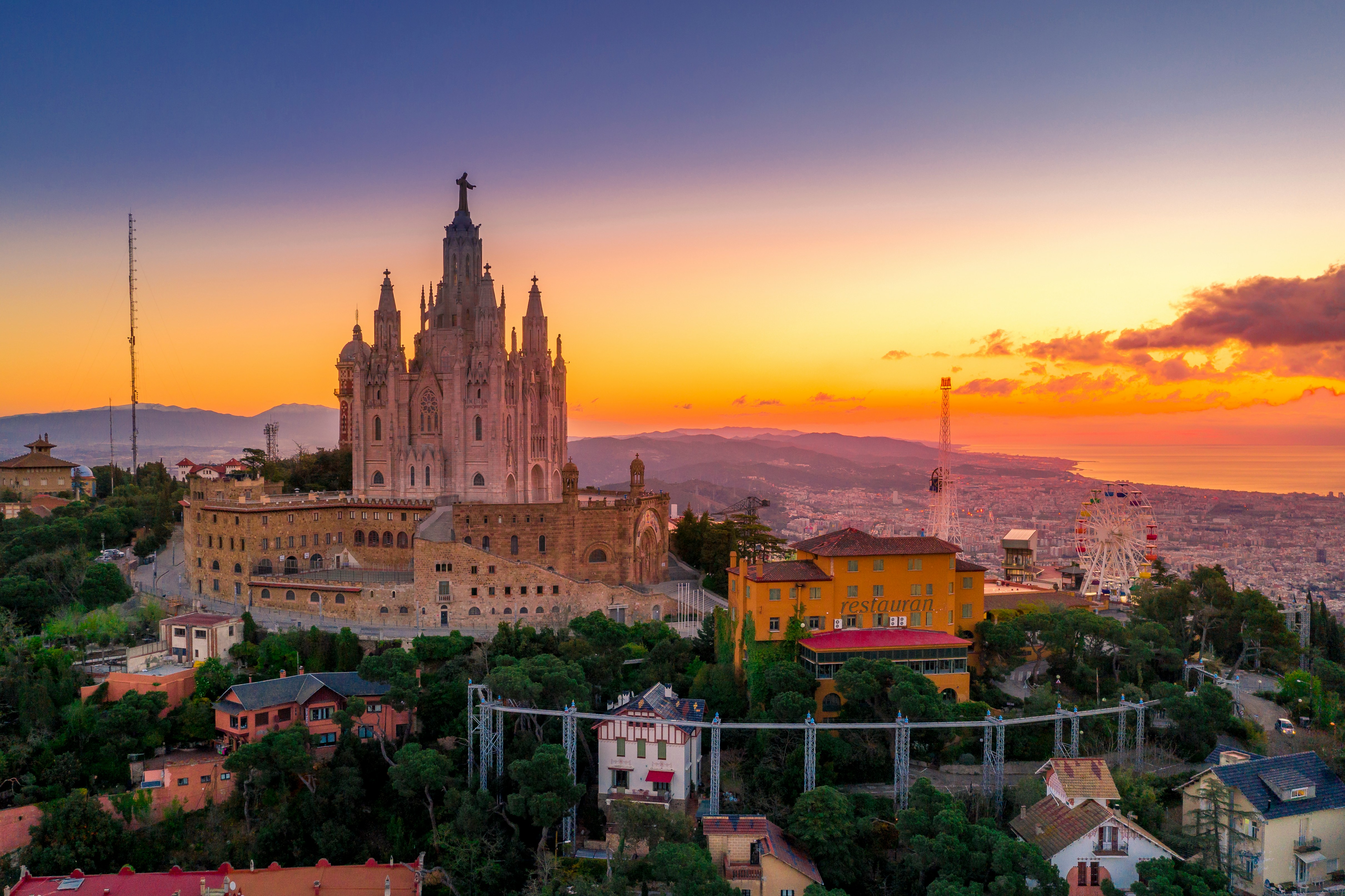 Barcelona at sunset from the Tibidabo hill, with the Sagrat Cor church silhouetted against an orange sky and the city spreading toward the sea below